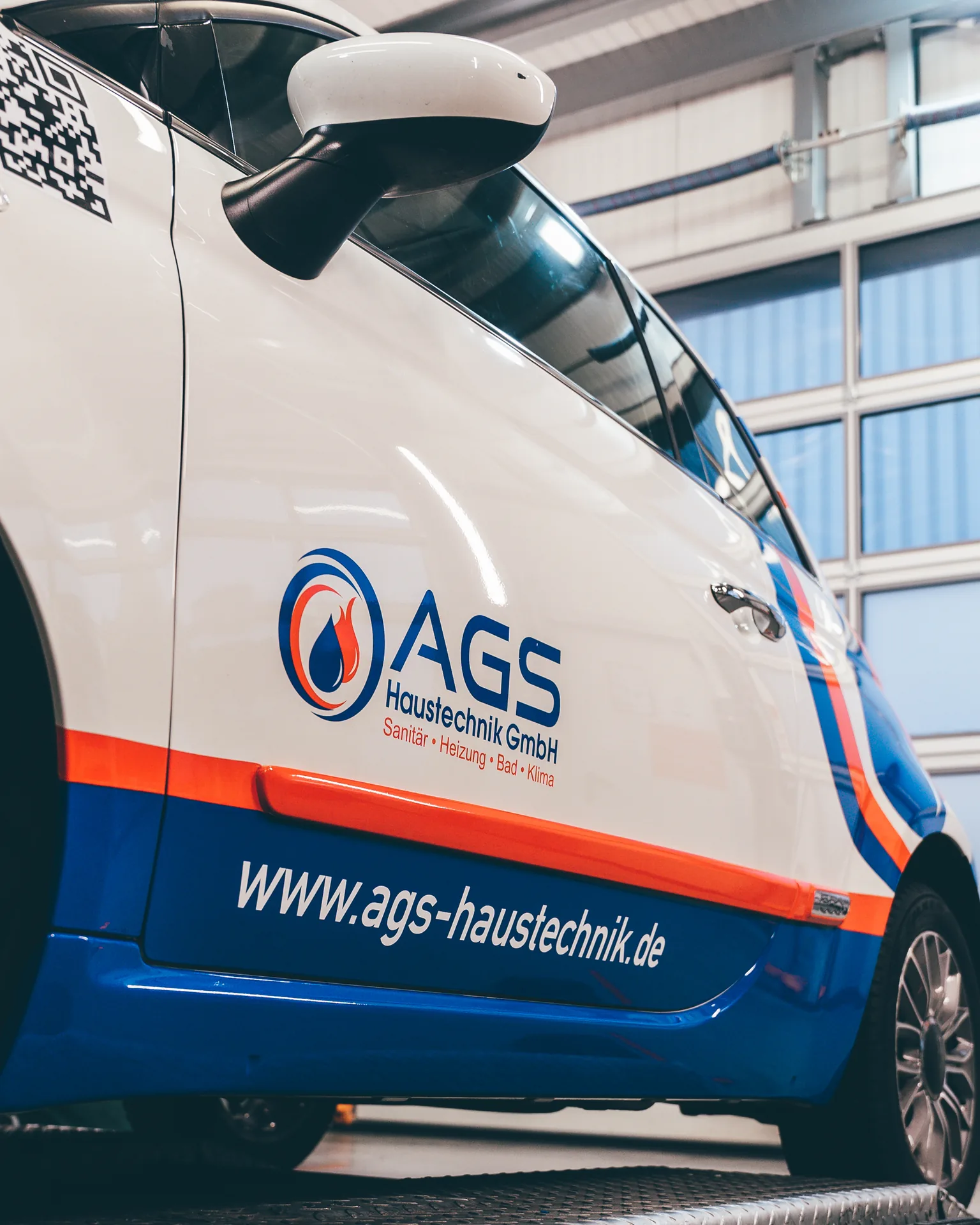 Beschrifteter weißer Transporter mit Firmenlogo, Web-Adresse und blau-orangefarbener Folie in der Werkstatthalle